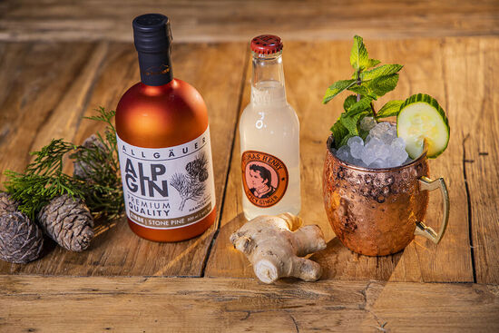 Serviervorschlag: Die kupferne Flasche AlpGin Zirbe steht neben einer Flasche Thomas Henry Spicy Ginger und einem kupfernen Cocktailbecher, der garniert ist mit Rosmarin und Gurke
